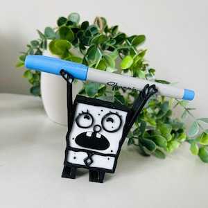 May include: A black and white 3D printed figurine of a cartoon character with a wide open mouth and large eyes. The character is holding a blue and white Sharpie marker above its head.