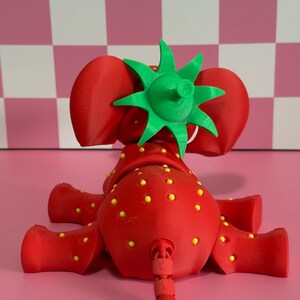May include: A whimsical red strawberry elephant figurine with yellow seed details and a green leafy top. The toy is 3D-printed and sits on a pink surface. The elephant has a green tail and large red ears.