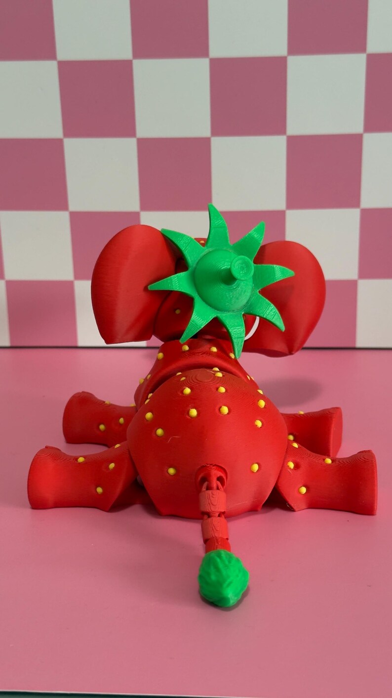 May include: A whimsical red strawberry elephant figurine with yellow seed details and a green leafy top. The toy is 3D-printed and sits on a pink surface. The elephant has a green tail and large red ears.