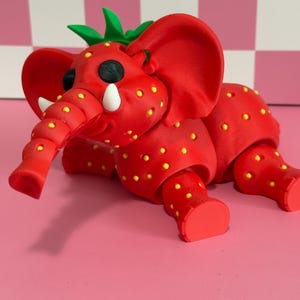 May include: A whimsical red elephant sculpture with a strawberry design. The elephant has yellow seeds, white tusks, and green leaves on its head. It is set against a pink background.
