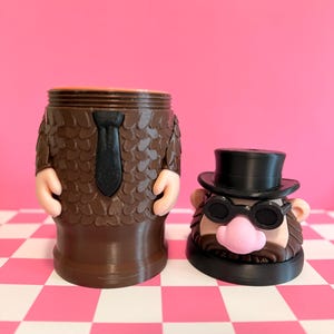 May include: A brown novelty container shaped like a man in a suit, with a black tie and arms. Beside it is a head with a top hat, glasses, and a pink nose. The background is pink and white checkered.