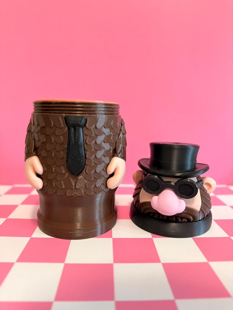 May include: A brown novelty container shaped like a man in a suit, with a black tie and arms. Beside it is a head with a top hat, glasses, and a pink nose. The background is pink and white checkered.