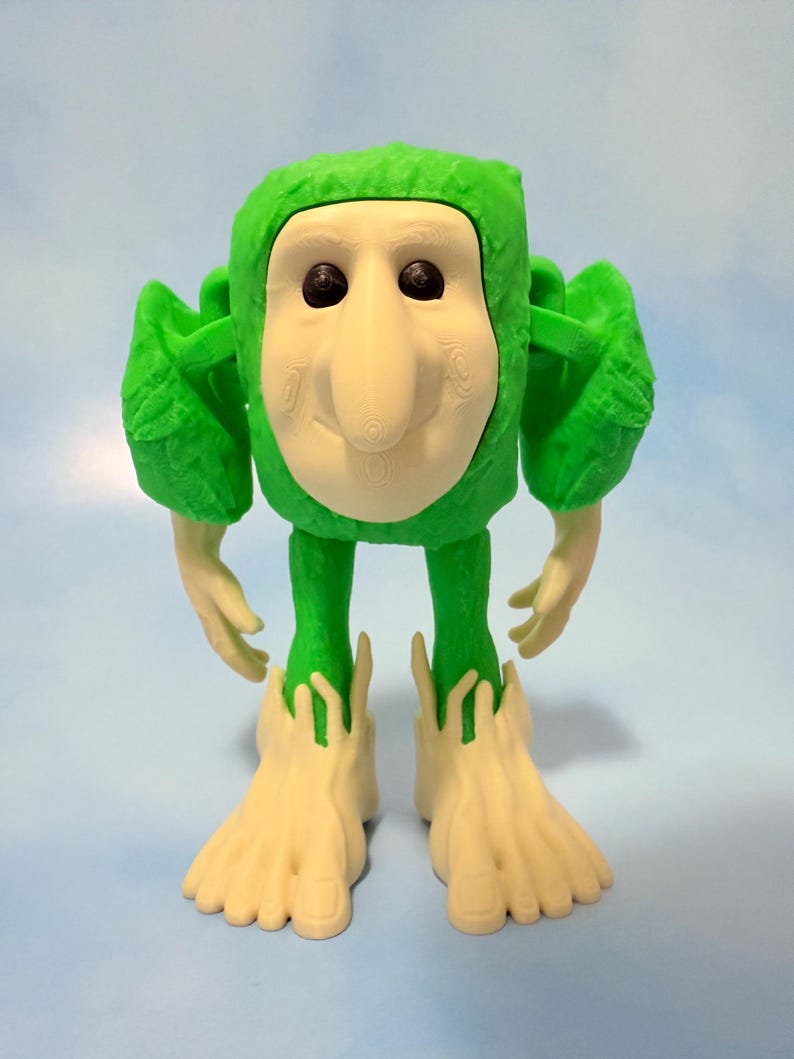 Op de afbeelding: Een grillig groen en gebroken witte beeldje met een uniek ontwerp. De figuur heeft een groene kop en lichaam, met gebroken witte gelaatstrekken, handen en voeten. De grote voeten en handen van de figuur zijn een opvallend kenmerk.