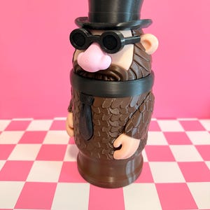May include: A novelty figurine resembling a distinguished gentleman, featuring a black top hat, round glasses, and a brown beard. The figure has a pink nose and tan ears, with a brown suit and a black tie. The figurine is standing on a pink and white checkered surface.