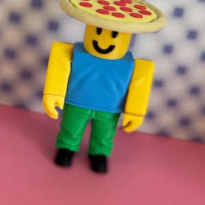May include: A yellow Roblox figure wearing a pizza hat, blue top, and green trousers. The pizza hat has a yellow crust and red pepperoni. The figure is standing on a pink surface.