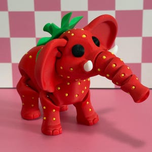 May include: A whimsical red elephant figurine with a strawberry design. The elephant has yellow seeds, green leaves, and white tusks. The background is a pink and white checkered pattern.