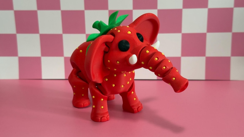 May include: A whimsical red elephant figurine with a strawberry design. The elephant has yellow seeds, green leaves, and white tusks. The background is a pink and white checkered pattern.