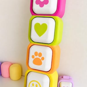 May include: A vertical stack of four square white buttons with colorful silicone frames. The top button has a pink flower, the second a lime green heart, the third an orange paw print, and the bottom a yellow smiley face. Additional small colorful items are nearby.