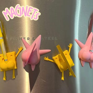 May include: Four 3D printed magnets in the shape of cartoon characters. Two are yellow and two are pink. The magnets are attached to a stainless steel refrigerator. The word "MAGNETS" is written in pink letters on the refrigerator.