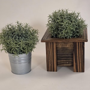 May include: Two decorative planters. One is a small, galvanized metal bucket filled with green foliage. The other is a dark brown, wooden planter with a square shape, also filled with green foliage. Both are on a white surface.