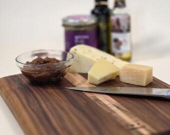 Puede incluir: Una tabla de embutidos de madera con una variedad de quesos, un pequeño cuenco de conservas y un cuchillo. Varias botellas de aceite y un tarro de conservas están en el fondo. La tabla es de madera oscura con un acento de madera más clara.