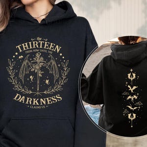 May include: Black hooded sweatshirt with a front graphic featuring the text "The Thirteen Darkness Claims Us" in gold, along with a sword, wings, and floral elements. The back of the sweatshirt has a vertical design with gold illustrations.