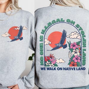 May include: Sport grey sweatshirt with a graphic of a flying eagle in front of a red sun and clouds. The back of the sweatshirt has the text "NO ONE IS ILLEGAL ON STOLEN LAND" and "WE WALK ON NATIVE LAND" with flowers.