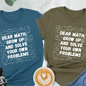 May include: Two t-shirts with a white graphic print of math symbols and the text "Dear Math, Grow Up and Solve Your Own Problems". The shirts are in heather deep teal and heather olive.