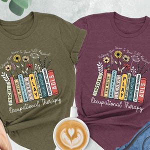 May include: Two t-shirts with a design of books stacked on top of each other with the words "Occupational Therapy" printed below. The books have different colored spines and the words on the spines are "Social", "Sensory Processing", "Fine Motor", "Cognition", "Learn", "Play", "Bilateral Coordination", "Visual Motor", "Executive Functioning", and "ADLS". The t-shirts are in heather maroon and heather olive.
