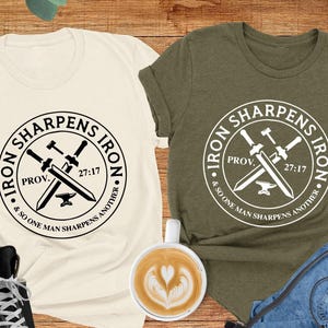 May include: Two t-shirts with a graphic design of a crossed sword and anvil with the text "Iron Sharpens Iron" and the Bible verse "Prov. 27:17 & So One Man Sharpens Another". The t-shirts are in natural and heather olive colors.