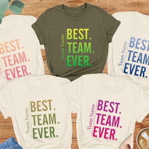 May include: Five t-shirts in different colors with the text "Best Team Ever" printed on them. The shirts are laid out on a wooden surface. The text is in a gradient of colors.