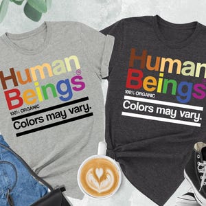 May include: Two t-shirts, one gray and one dark gray, each with the text "Human Beings" in colorful letters. Below the text is "100% ORGANIC" and "Colors may vary." A pair of jeans, a black purse, a cup of coffee, and a pair of black high-top sneakers are also visible.
