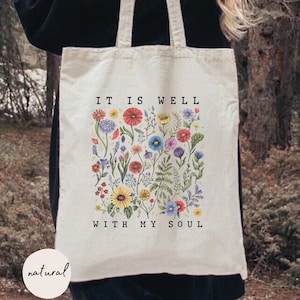 May include: Cream-colored tote bag with the text "IT IS WELL WITH MY SOUL" and a floral design. The design features colorful flowers and greenery. The bag has two handles and is being held by a person.