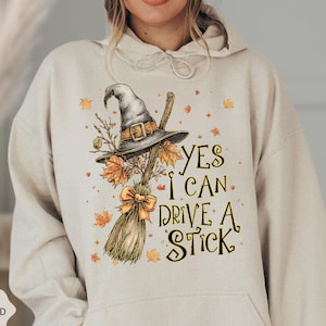 May include: Sand-colored hoodie with a witch hat, broom, and autumn leaves graphic. The text reads "YES I CAN DRIVE A STICK". The design includes a bow and decorative elements.