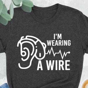 Pode incluir: Uma camiseta cinza escuro mesclada com um gráfico branco de uma orelha estilizada com uma linha de batimento cardíaco e o texto "I'm Wearing A Wire".