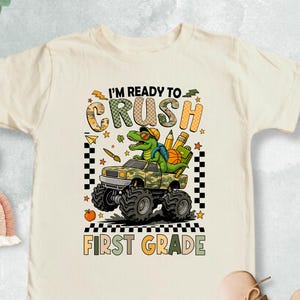 May include: Cream-colored toddler t-shirt with a colorful graphic. The graphic features a green dinosaur driving a monster truck with the text "I'M READY TO CRUSH FIRST GRADE". The shirt has short sleeves.