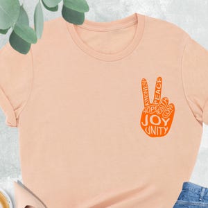 May include: A heather peach t-shirt with a peace sign graphic in orange. The peace sign includes the words "Kindness, Peace, Hope, Faith, Love, Joy, Unity." The shirt is paired with light wash blue jeans and nude flats.
