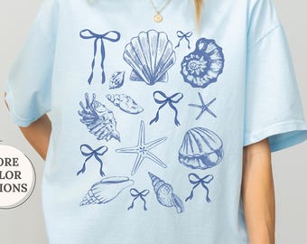 May include: Light blue t-shirt with a seashell and bow graphic. The shirt features various seashell illustrations and bows in a darker blue color. The word "CHAMBRAY" is printed at the top right. The words "MORE COLOR OPTIONS" are printed on a white circle.
