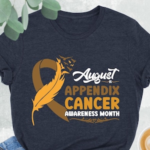 May include: A heather navy blue t-shirt with a brown ribbon and a yellow feather. The text on the shirt reads "August is Appendix Cancer Awareness Month".