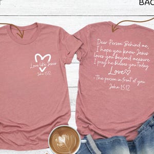 May include: Heather mauve t-shirt with white text and a heart design. The front of the shirt says "Love like Jesus" and "John 15:12". The back of the shirt has a longer message. The shirt is paired with jeans and white sneakers.