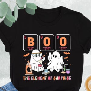 May include: Black t-shirt with a Halloween-themed graphic. The design features the word "BOO" spelled out using the periodic table symbols for Boron and Oxygen, along with two ghost characters and the text "THE ELEMENT OF SURPRISE".