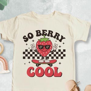 May include: Cream-colored toddler t-shirt with a graphic of a strawberry wearing sunglasses and riding a skateboard. The text on the shirt reads "SO BERRY COOL". The design includes a checkered pattern and peace signs. The shirt is paired with small shoes.