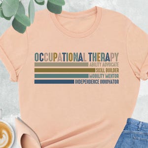 May include: A peach-colored t-shirt with the words "OCCUPATIONAL THERAPY" in blue and brown. Below the text are horizontal stripes and the phrases "ABILITY ADVOCATE", "SKILL BUILDER", "MOBILITY MENTOR", and "INDEPENDENCE INNOVATOR". The shirt is paired with blue jeans and nude-colored flats.