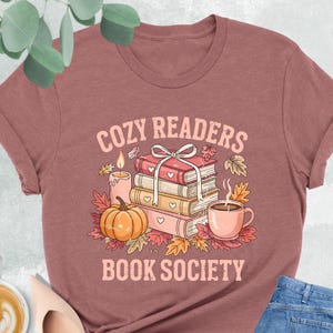 May include: Heather mauve t-shirt with the text "COZY READERS BOOK SOCIETY". The graphic includes a stack of books, a pumpkin, a cup of coffee, and autumn leaves. A pair of light pink shoes, a cup of coffee, and blue jeans are also in the image.