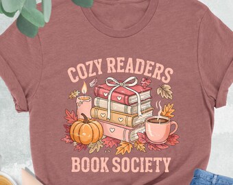 May include: Heather mauve t-shirt with the text "COZY READERS BOOK SOCIETY". The graphic includes a stack of books, a pumpkin, a cup of coffee, and autumn leaves. A pair of light pink shoes, a cup of coffee, and blue jeans are also in the image.