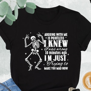 May include: Black t-shirt with a skeleton graphic and the text "ARGUING WITH ME IS POINTLESS I KNEW I was wrong 10 minutes ago I'M JUST Trying to MAKE YOU MAD NOW". The shirt is paired with blue jeans, a black purse, and black and white sneakers.