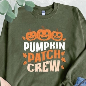 May include: Military green sweatshirt with the text "PUMPKIN PATCH CREW" in white, orange, and cream. The design includes three orange jack-o'-lanterns and fall leaves. The sweatshirt is a crew neck style.