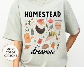 May include: A light green t-shirt with the word "HOMESTEAD" in black block letters. Below the word is the word "dreamin'" in a cursive font. The shirt features a colorful graphic of gardening and farm-related images.