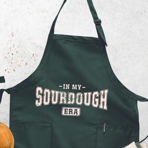 May include: Dark green apron with the text "-IN MY- SOURDOUGH ERA" in white. The apron has two pockets and a pen holder. A wooden spoon, a tea towel, and lit candles are in the background.