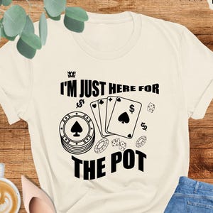 May include: Cream-colored t-shirt with the text "I'M JUST HERE FOR THE POT" in black. The design includes poker chips, playing cards, and dollar signs. The shirt is paired with blue jeans and pink shoes.