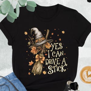 Pode incluir: Camiseta preta com um chap&eacute;u de bruxa, vassoura e folhas de outono. O texto diz "YES I CAN DRIVE A STICK". O conjunto inclui jeans azuis, uma bolsa preta e t&ecirc;nis de cano alto pretos.