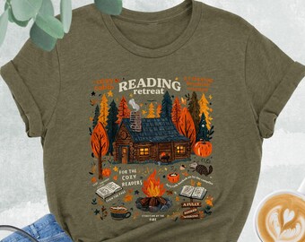 May include: A heather olive t-shirt featuring a graphic of a log cabin in the woods with a campfire, books, and the text Reading Retreat for the Cozy Readers. The shirt is paired with blue denim jeans, a black crossbody bag, and black sneakers.