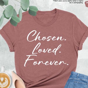 Pode incluir: Uma t-shirt em tom de malva mesclado com as palavras "Chosen. Loved. Forever." em escrita branca. A t-shirt é combinada com jeans azuis e sapatilhas bege. Uma chávena de café com arte latte também é visível.