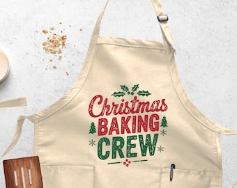 May include: A cream-colored apron with the text "Christmas BAKING CREW" in red and green. The apron has two pockets and a pen holder. The design includes holly leaves, a Christmas tree, and snowflakes.