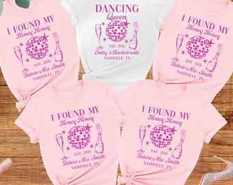 May include: A collection of five short-sleeved t-shirts in white and light pink. The shirts feature a pink graphic print with a disco ball, champagne glass, and bottle, along with the text I FOUND MY Honey Honey, DANCING QUEEN, and NASHVILLE, TN.