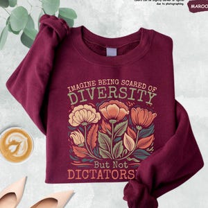 May include: Maroon sweatshirt with the text "IMAGINE BEING SCARED OF DIVERSITY But Not DICTATORS" and a floral design in shades of orange, beige, and green. The sweatshirt is displayed on a flat surface with a cup of coffee and a pair of shoes.