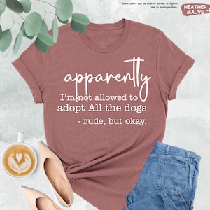 May include: Heather mauve t-shirt with the text "apparently I'm not allowed to adopt All the dogs - rude, but okay." The shirt is paired with blue jeans and nude flats. A cup of coffee with latte art is also visible.