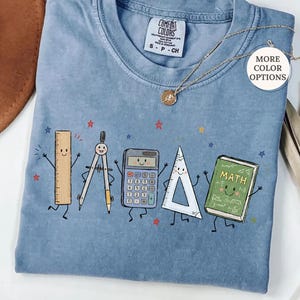 May include: A blue jean colored t-shirt featuring a graphic print of cartoon school supplies including a ruler, compass, calculator, triangle, and a green math book with arms and legs. The shirt has a crew neckline and a Comfort Colors brand label.