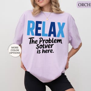 May include: A light purple t-shirt with the word "RELAX" in blue block letters. Below, the text reads "The Problem Solver is here" in black. The shirt is part of a clothing collection.