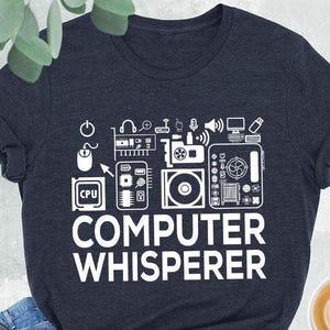 May include: Heather navy t-shirt with the text "COMPUTER WHISPERER" in white. The design includes computer-related icons. The shirt is paired with blue jeans, a black purse, and black high-top sneakers.